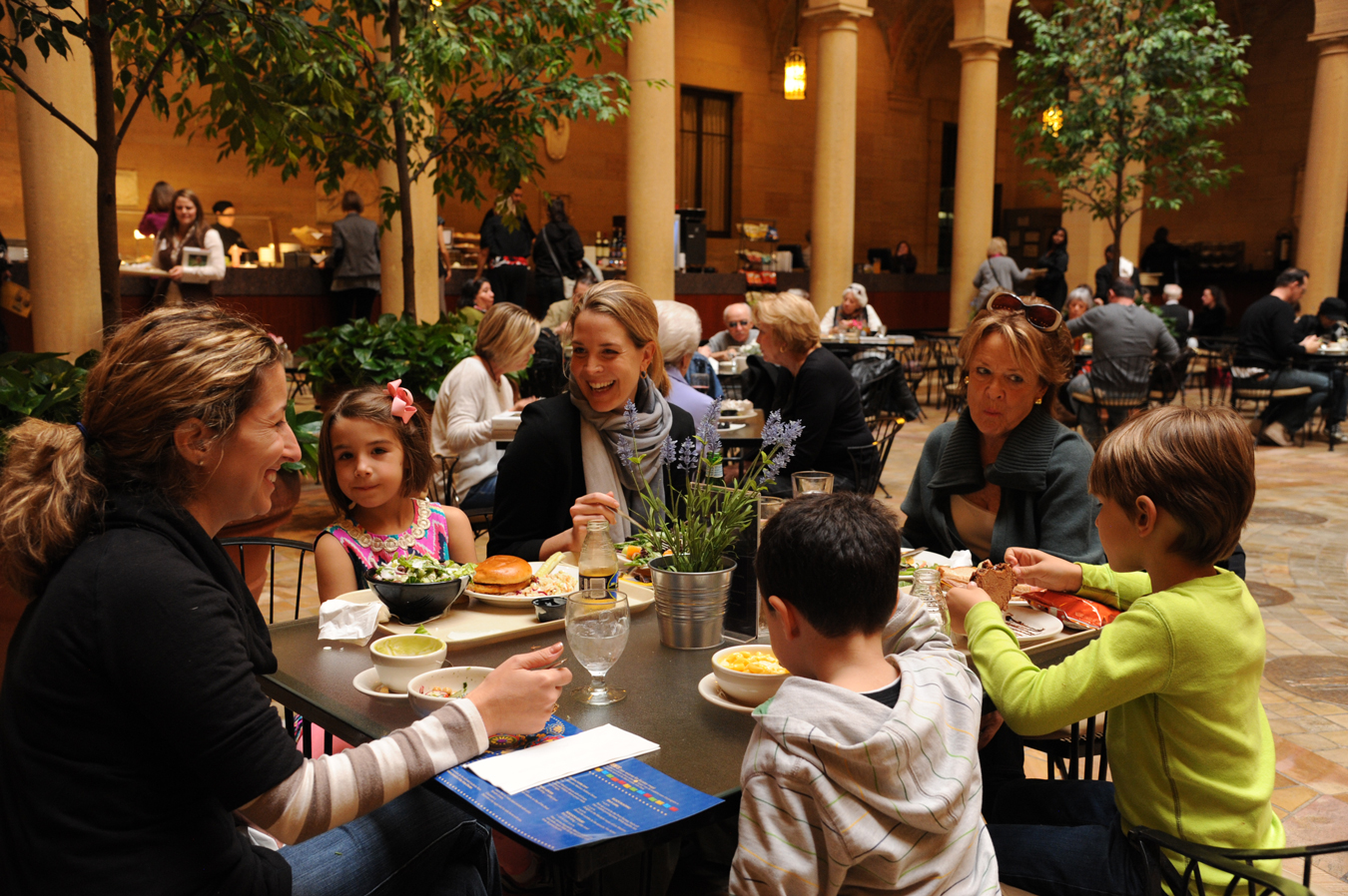 Group of three smiling women and three children enjoying lunch at a table in busy Rozzelle Court Restaurant.