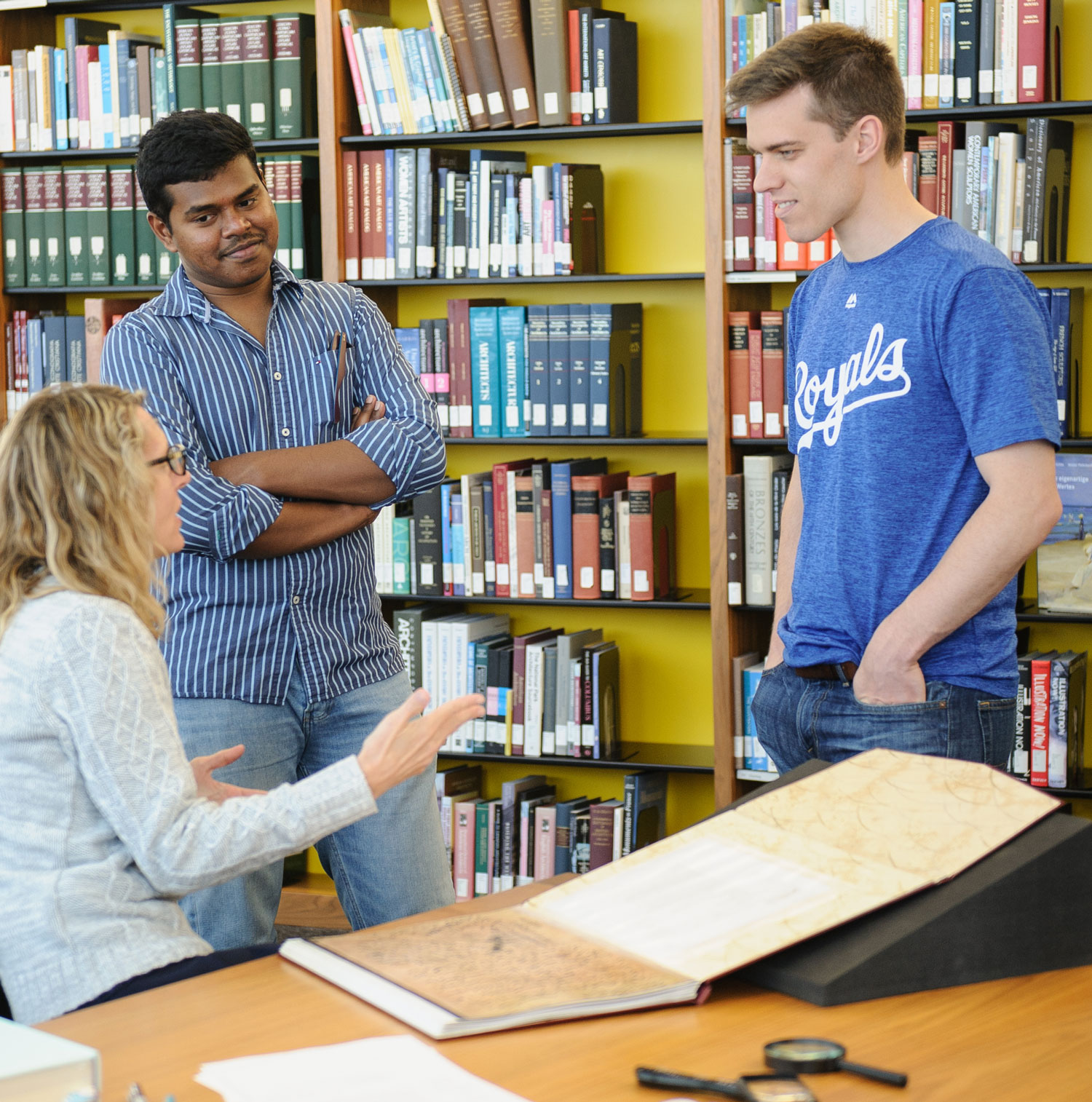 Librarian sitting at a table speaking with two people standing in front of shelves of books.