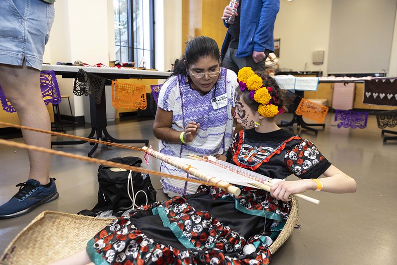 A young child dressed in a colorful skull dress weaves with the help of an instructor.