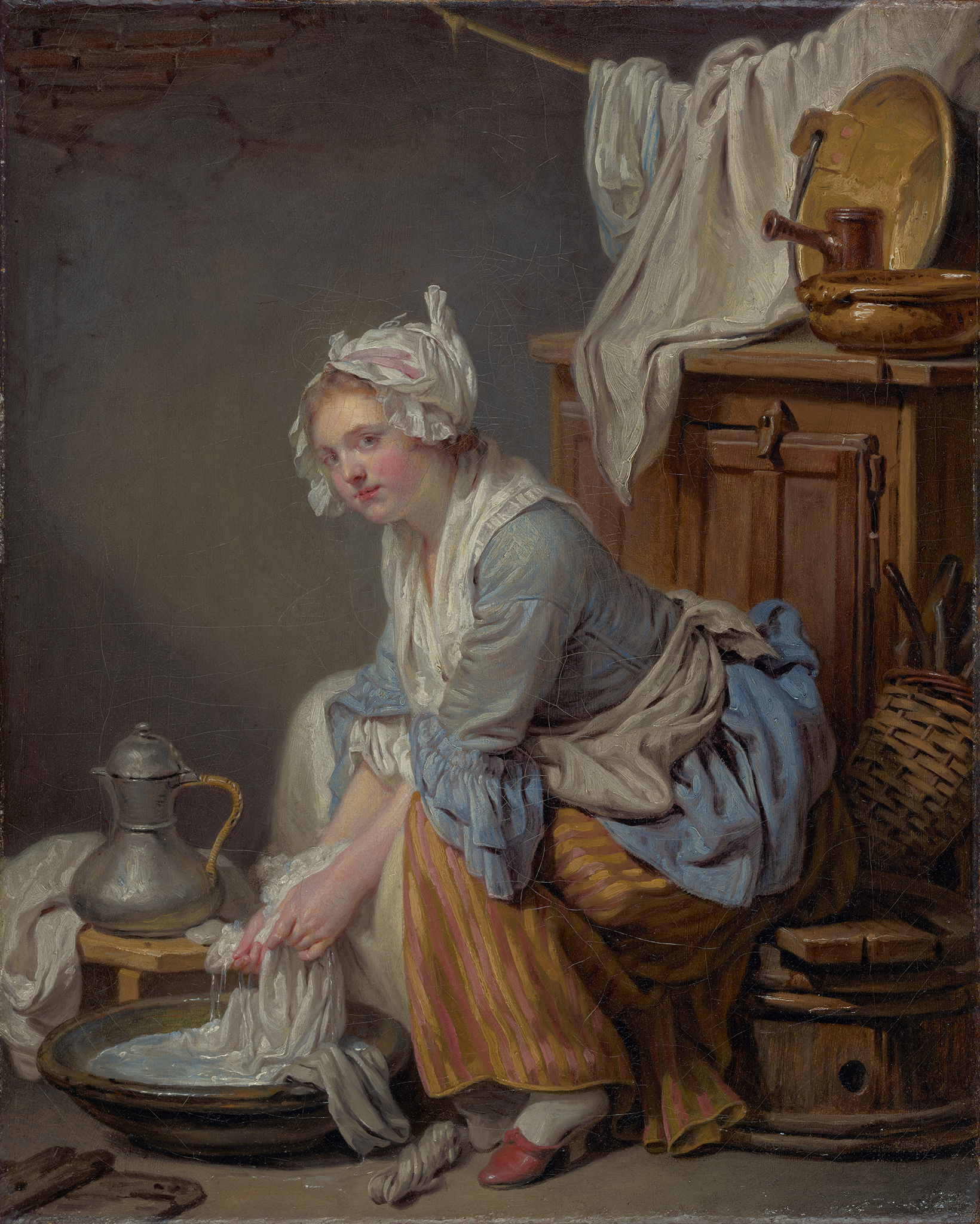 A painting of a woman wearing a light blue blouse and a striped orange and yellow dress.  She sits on a brown wooden barrel while washing clothes and fabrics in a bowl filled with water on the floor in front of her. Behind her is a wooden cabinet with various wooden tools resting on and around it. Above the dresser, there is a clothesline white a white fabric hung over it. In the background, the grayish blue paint on the wall has been chipped off showing a layer of bricks.