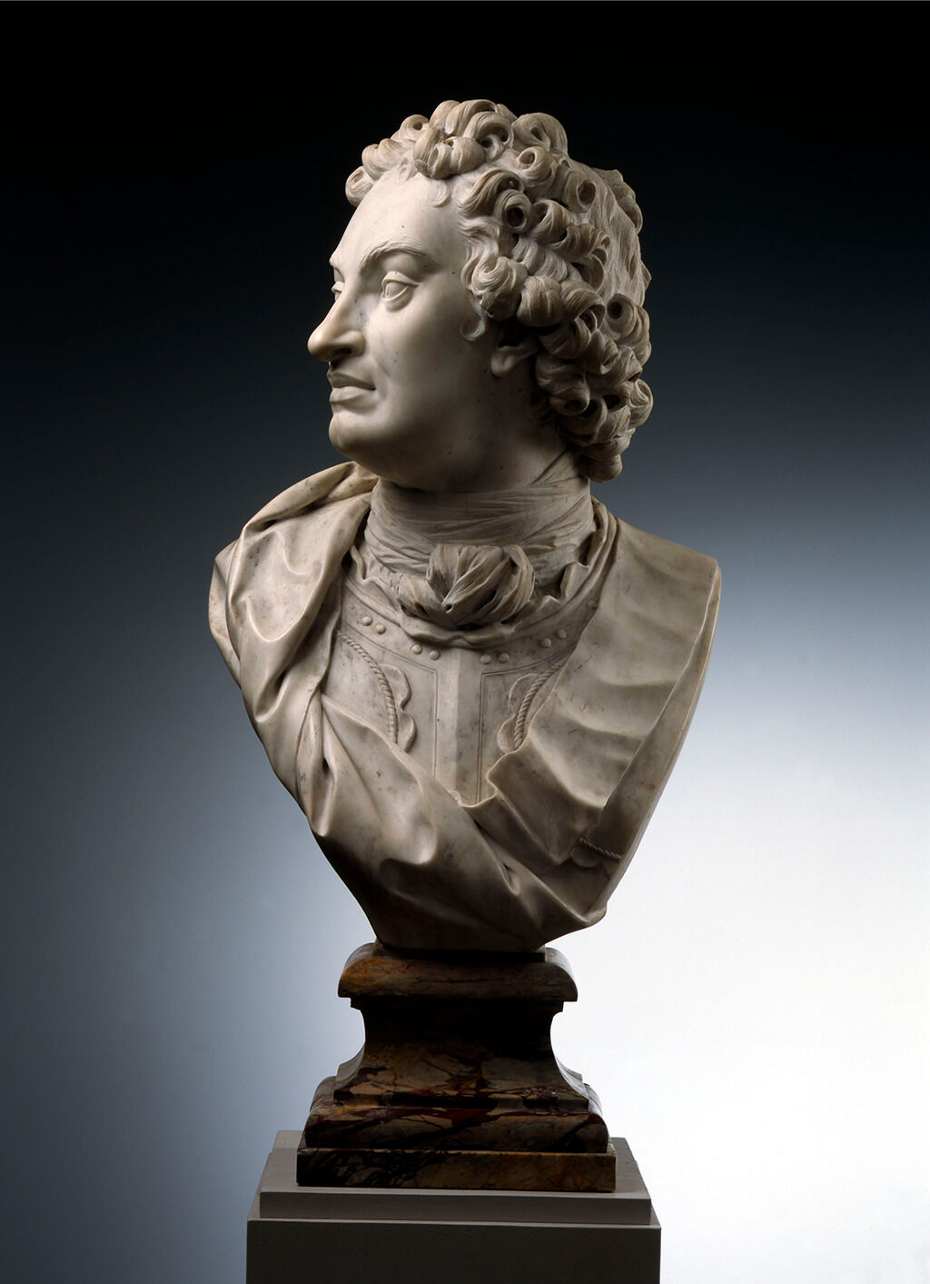 A photograph of a white sculpture of the side of a man’s face. The figure also has very curly hair sculped in along with a large nose. Around in his neck is a necktie, and under it seems to be a metal chest plate.