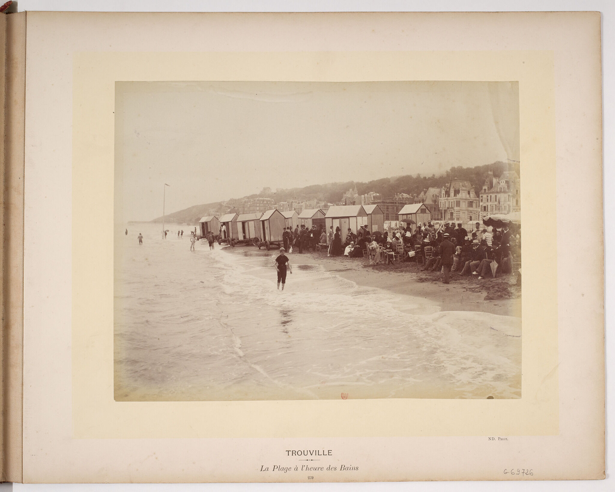 A painting depicting three large ships with colorful flags attached to the halyard, are docked in the harbor. In the water we see small boats filled with passengers and also people swimming. In the background, there are rows of buildings.