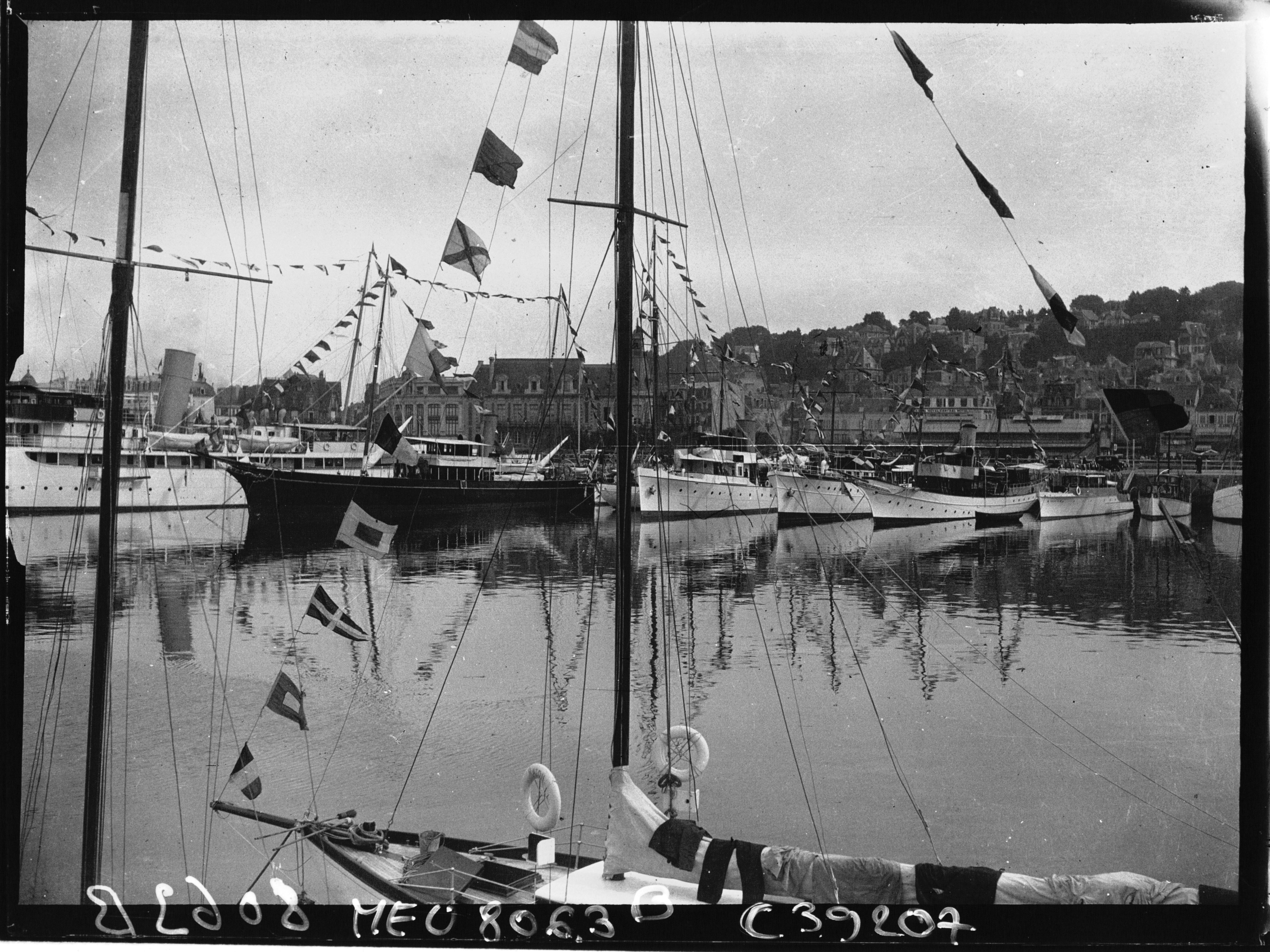 A photograph of a dock line with black and white sailboats. There are also dozens of small flags connected to the sailboat’s mass. The background consists of a large town filled with buildings and hills.