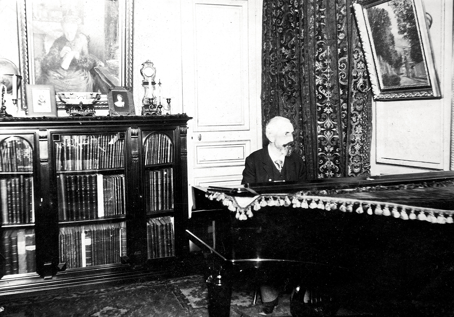 A black and white photograph of a man sitting behind a large piano. To his right is a long bookshelf with a framed painting above it.