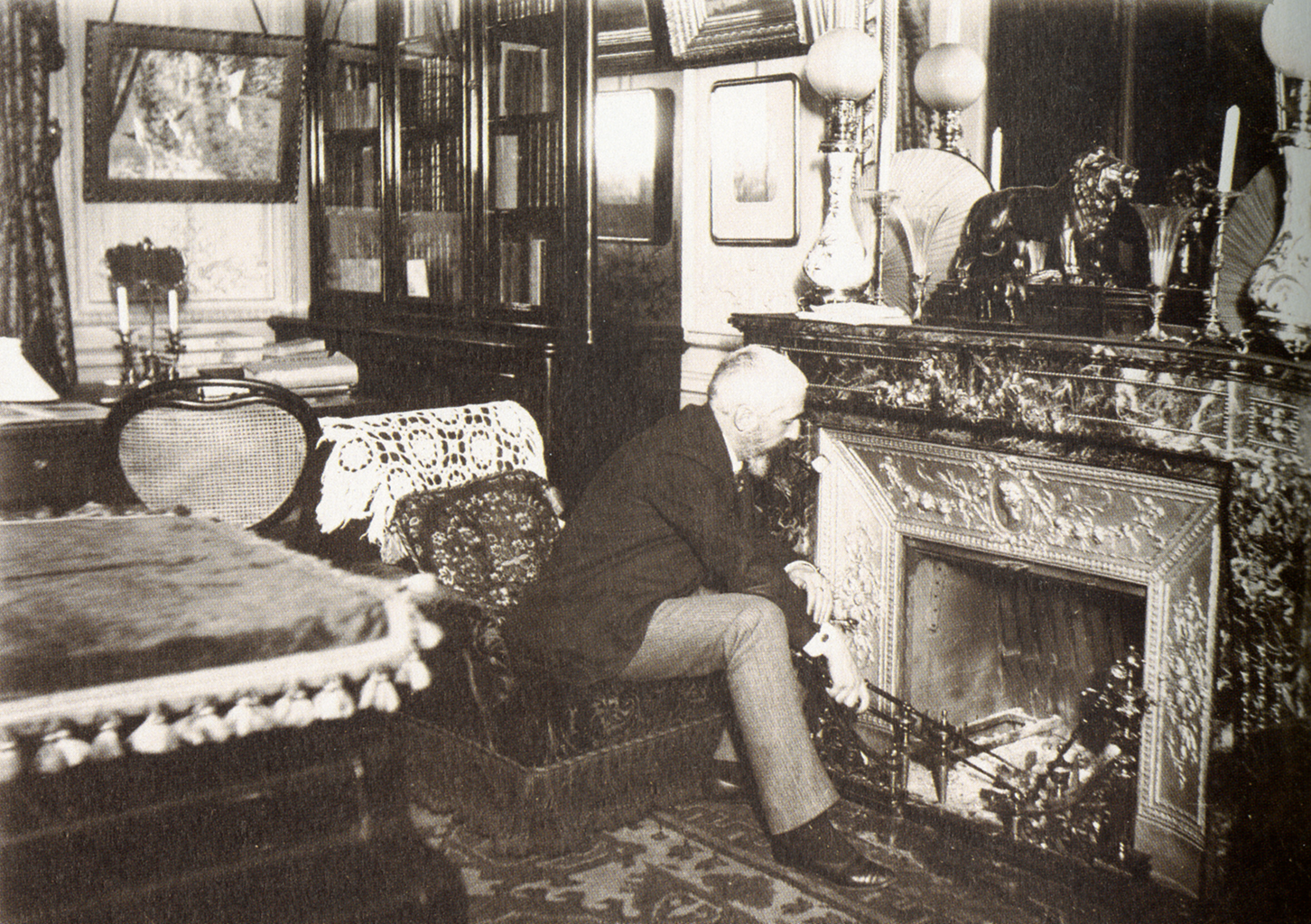 A black and white photograph of a man facing an ornamental fireplace. In the background there is a large bookshelf with other paintings hanging around it.