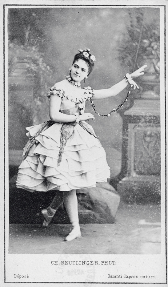 A black and white photograph of a woman wearing a white dress. She has her legs crossed and her left arm pointing upward at an angle. Along her dress are various flowers and vines. The woman also has some pinned on her hair.