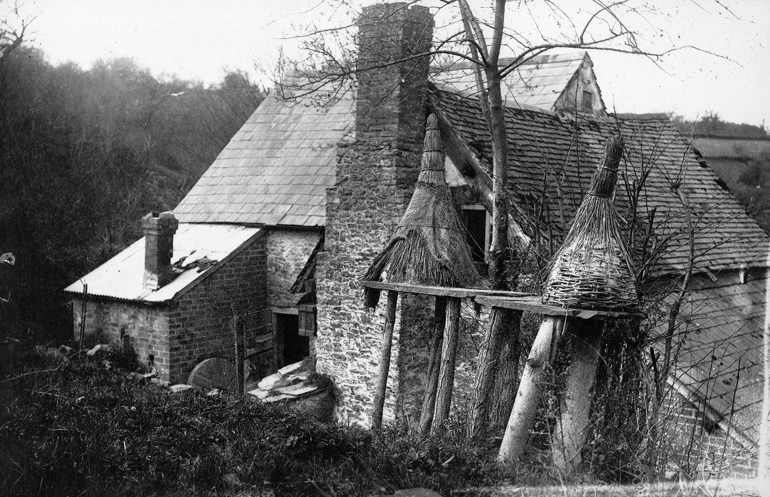 A black and white photograph of a house with a large, sloped roof and a tall brick chimney. There are also two straw beehives near it.