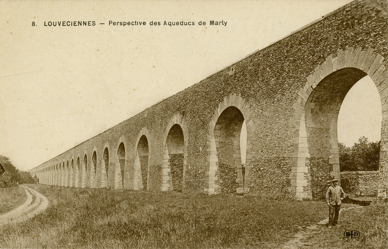 A sepia-toned photograph of a man standing in a grass field next to a towering wall made of cobblestone that has dozens of archways.