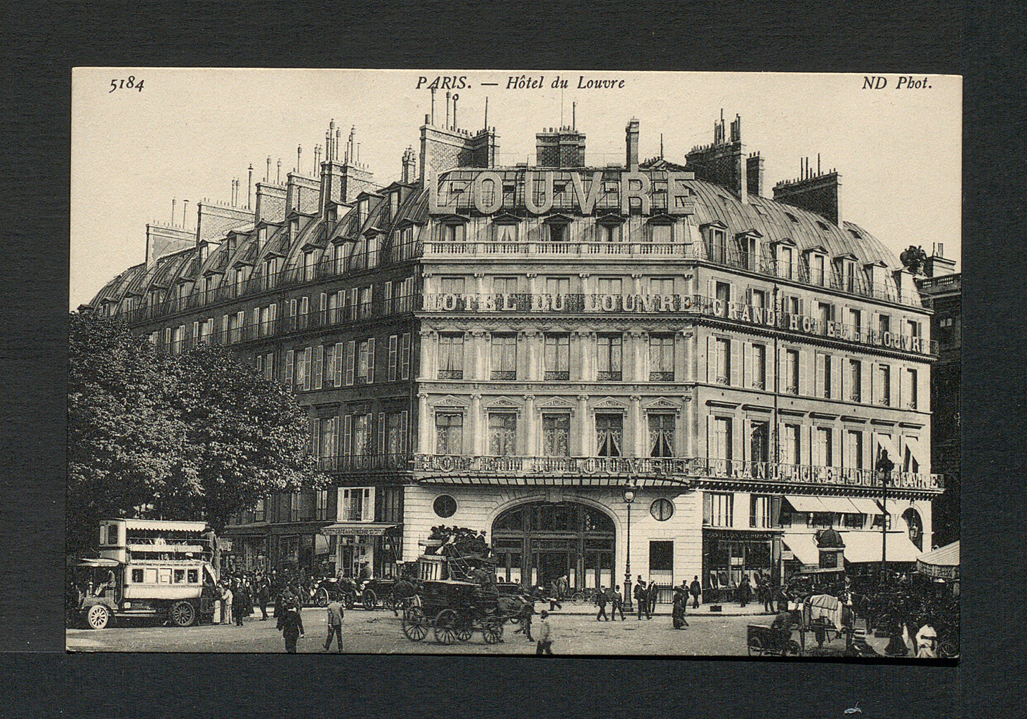 A black and white photograph of a long building with dozens of windows. There are letters on the front of the building which spell out &ldquo;LOUVRE&rdquo; The doors to the building are arched and surrounded by people and carriages. At the top of the photograph the words &ldquo;5184&rdquo;, &ldquo;PARIS. - Hotel du Louvre&rdquo;, and &ldquo;ND Phot.&rdquo; are written.