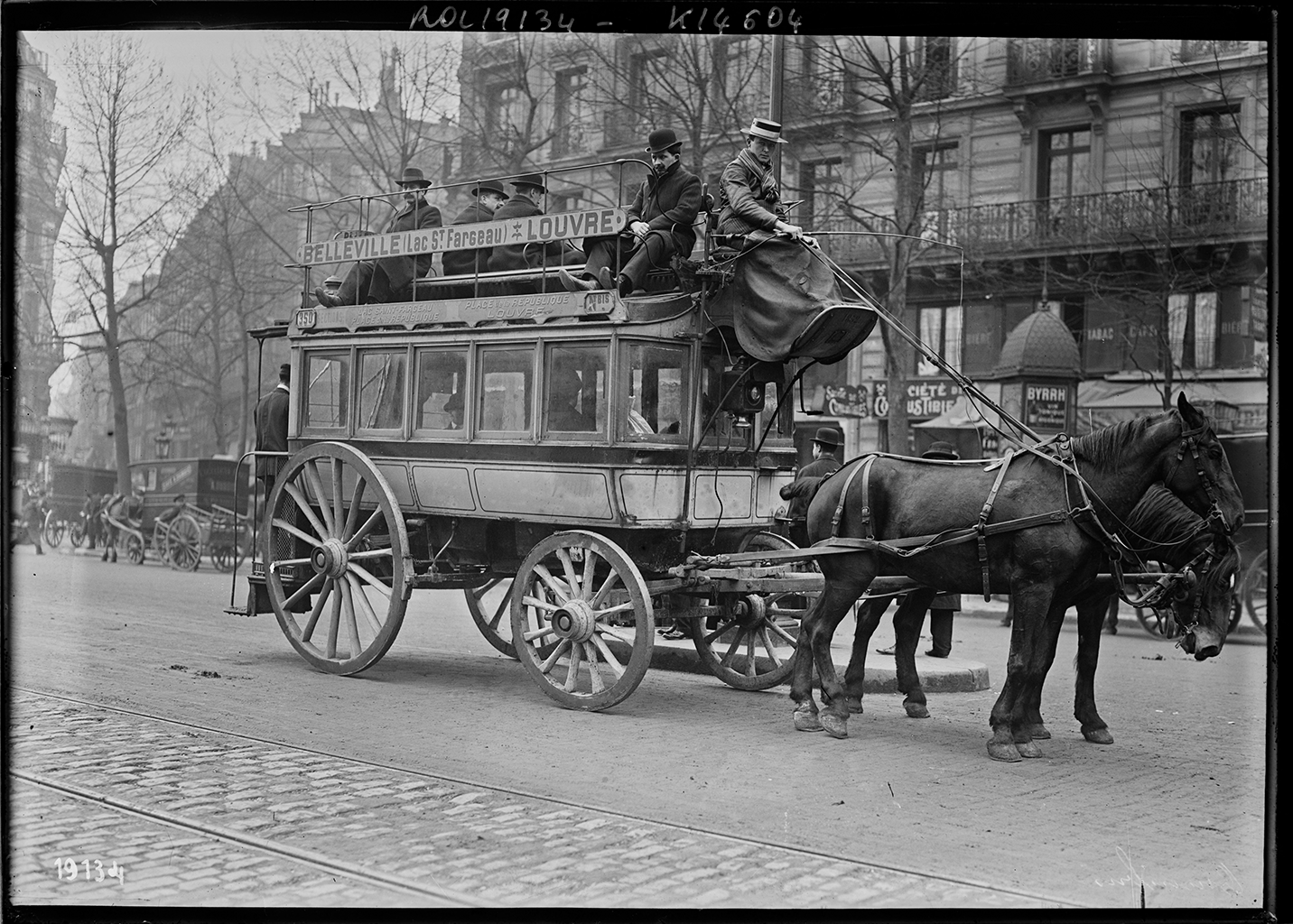 A black and white photograph of a light-colored carriage bring pulled by two dark colored horses on a road. The carriage has men sitting on the top of it while there appears to people sitting inside it as well. A man wearing a flat hat with a long gray dress or blanket over his legs. In the background, there is the façade of a building with windows and fences. There are also other horse carriages parked along the road.