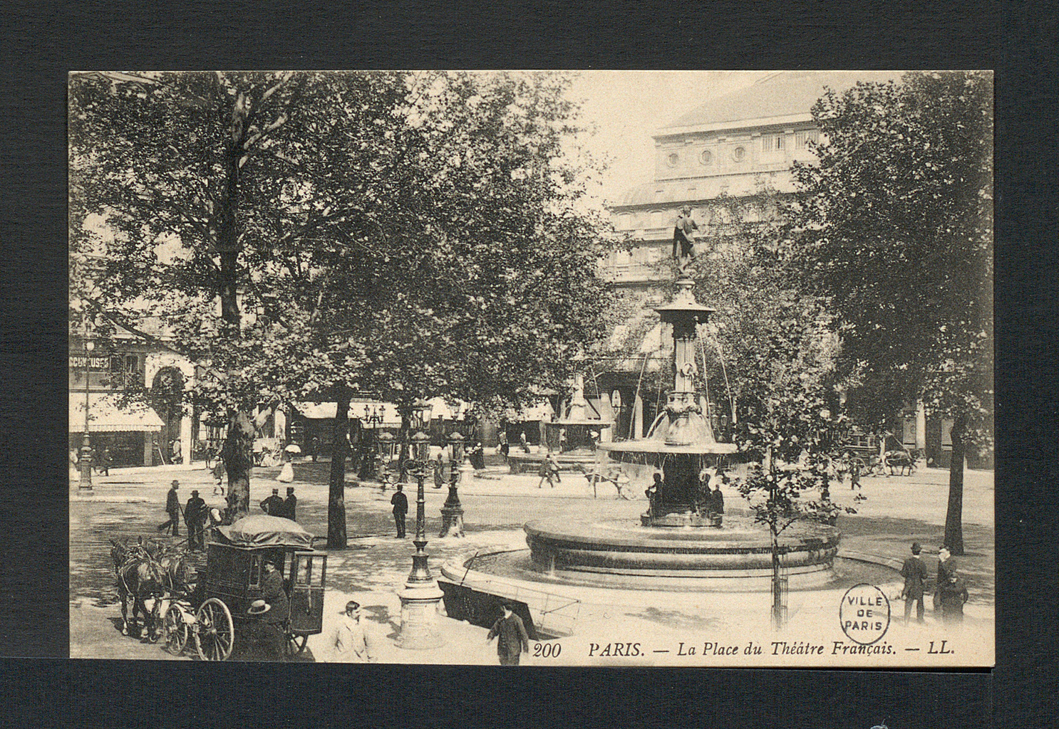 A black and white photograph of a round fountain surrounded by people and trees. In the background, there is a road with buildings that are aligned against it.