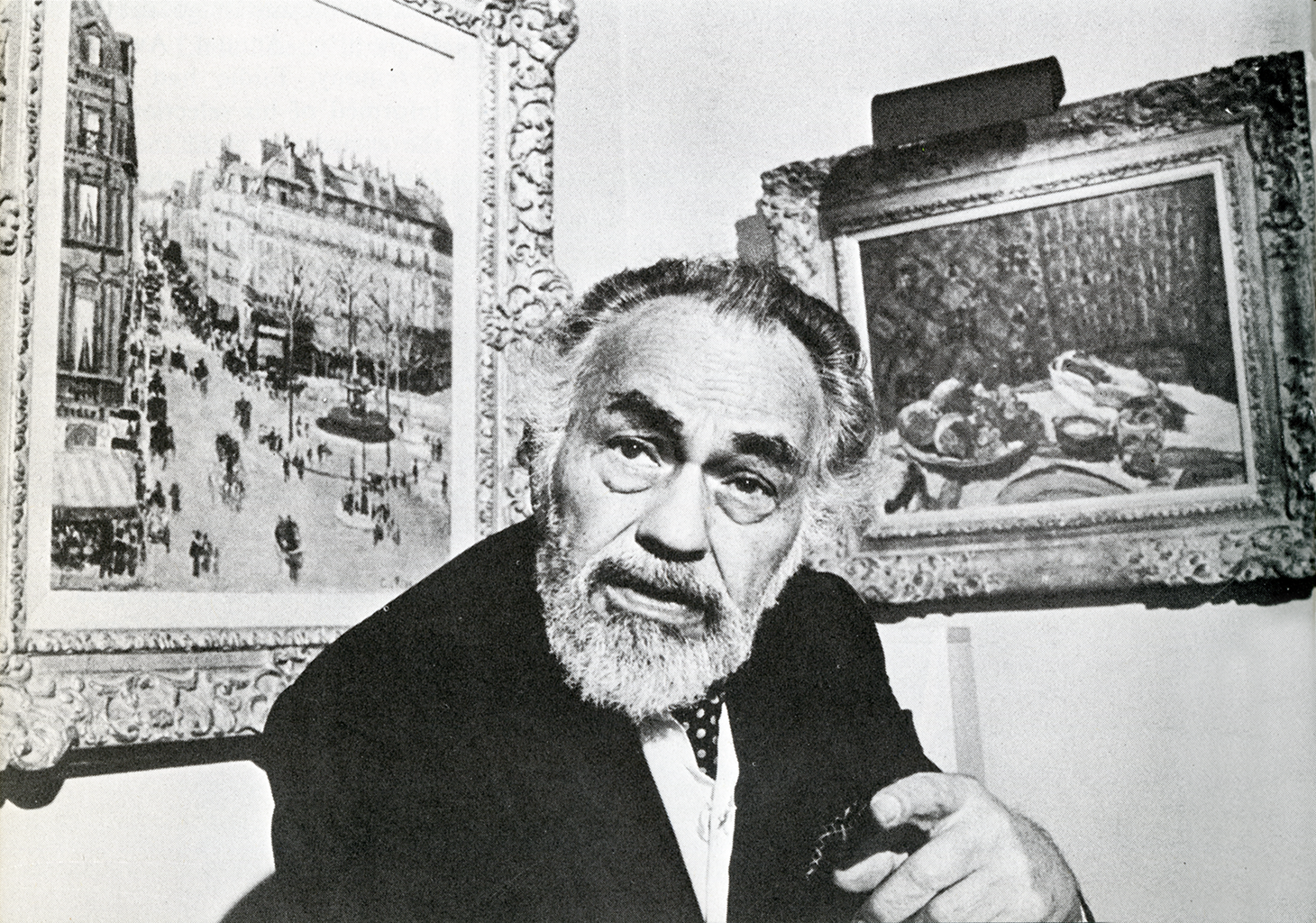 A black and white photograph of an older man with thick eyebrows and a gray beard. He wears a suit and points toward the camera. There are two impressionist-style paintings framed and hanging on the wall behind him.