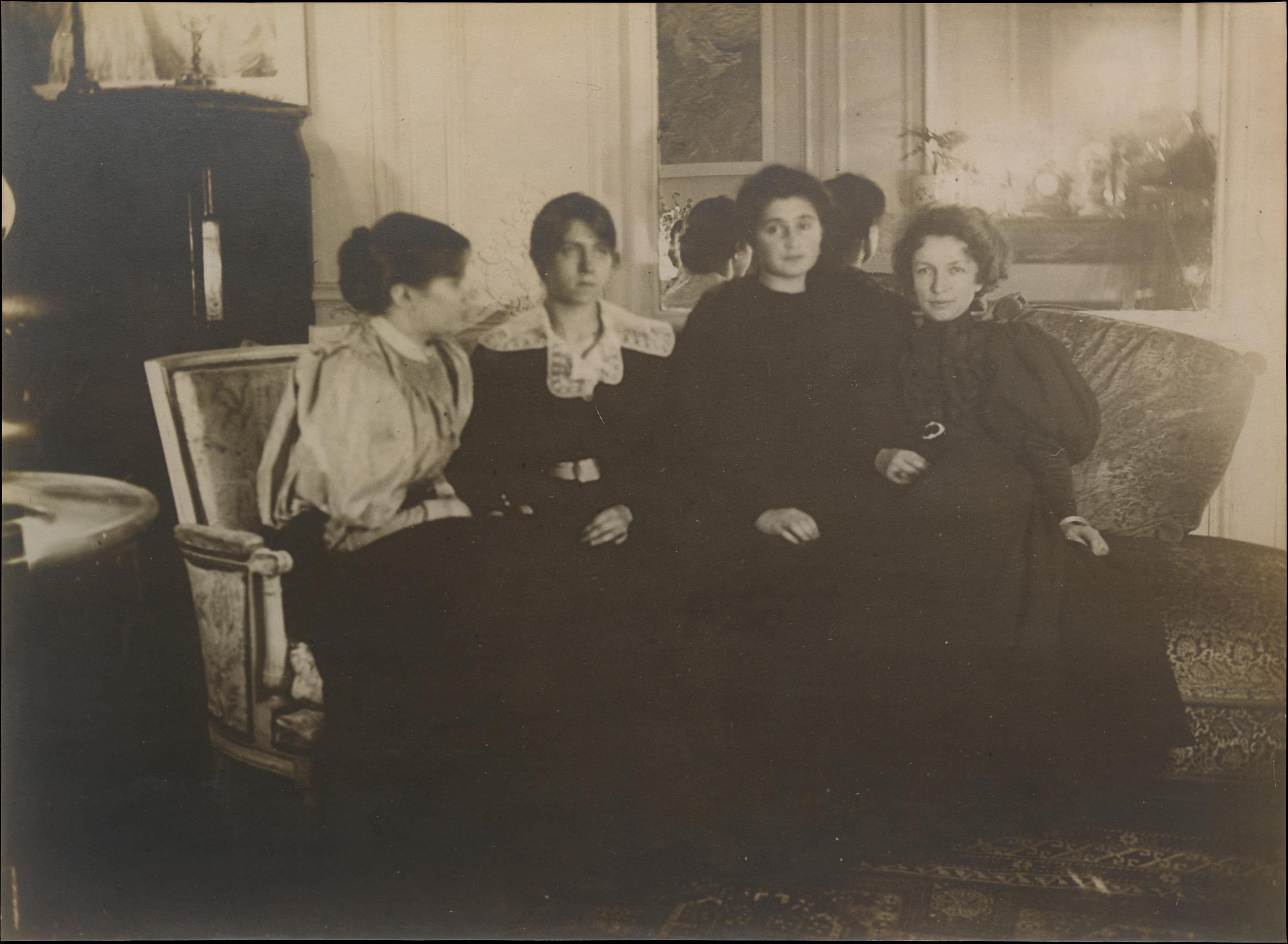 A black and white photography of four women wearing black or dark dresses sitting together on a coach. The background consists of a mirror on the wall and a cabinet to the left.