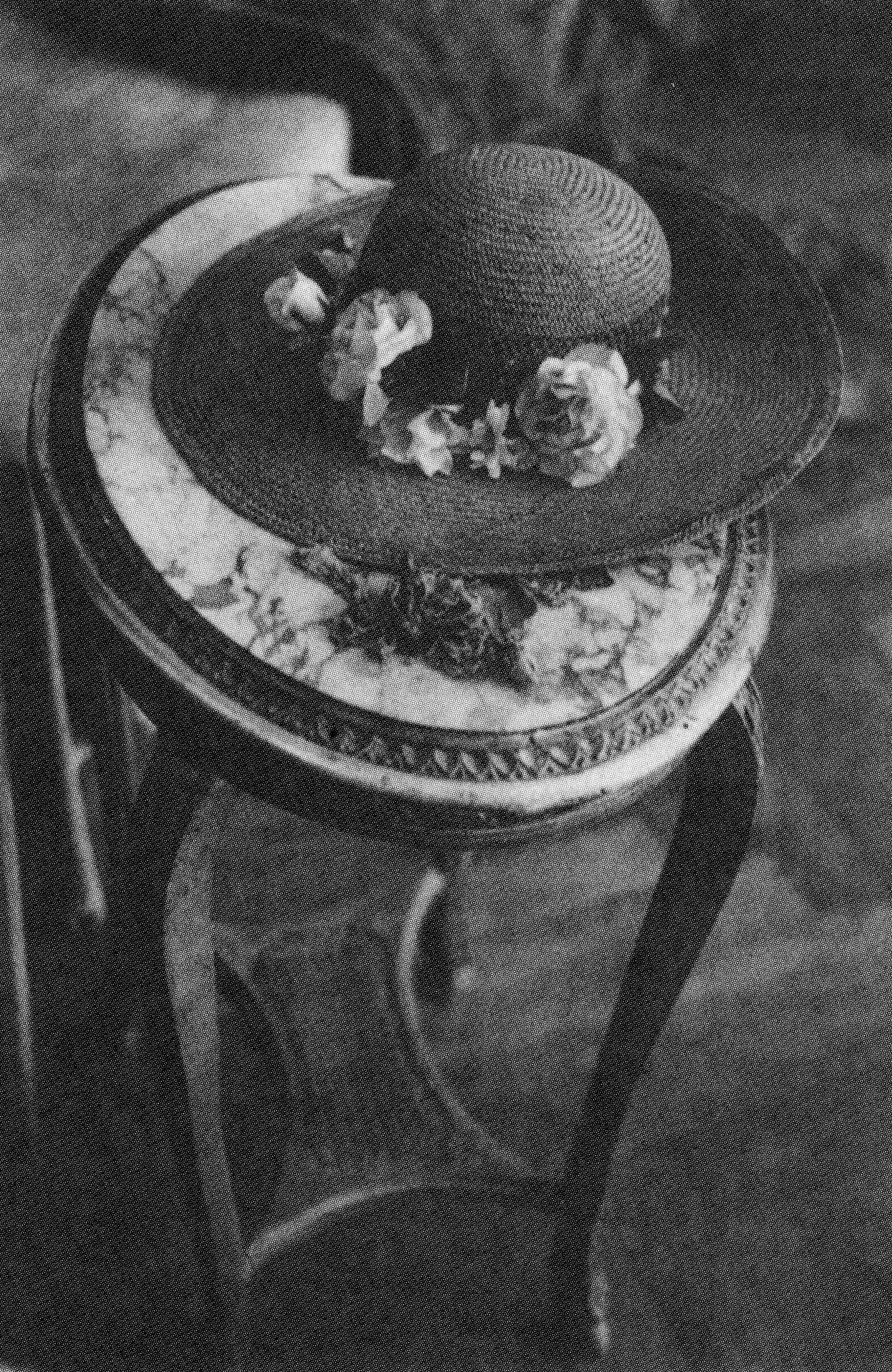 A black and white photograph of a straw hat with flowers on it set on an oval granite table.