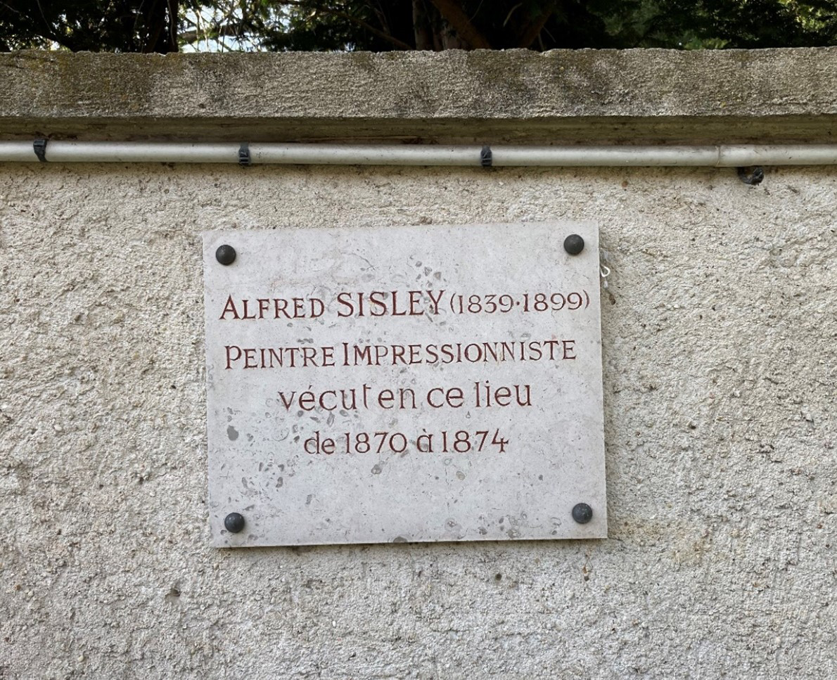 A photograph showing a textured gray stone wall with a metal plaque bolted onto the side of it. It states, &ldquo;ALFRED SISLEY (1839-1899) PEINTRE IMPRESSIONNISTE vécut en ce lieu de 1870 à 1874.