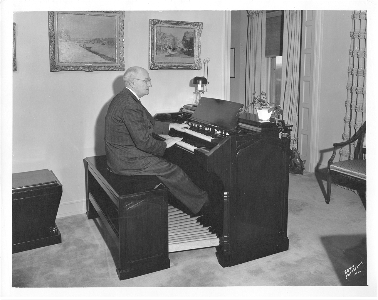 A black and white photograph of a man in a suit sitting on a bench behind an organ. His hands are resting on the keys of the instrument. Behind are two paintings which are hung along the wall.