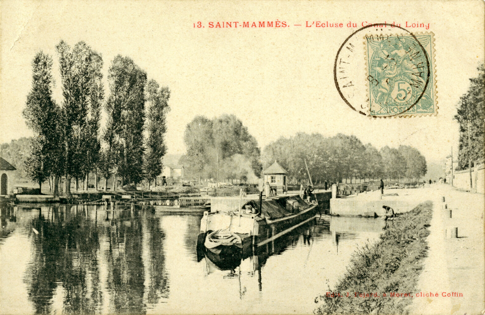 A black and white image of a dock on a river lined with trees and a walking trail. The path is near a small grass shore. A boat with boxes and tarps waits at the dock. On the image is a green stamp, and above it writes &ldquo;3. Saint-Mammès. - L’Ecluse du Canal du Loing&rdquo;