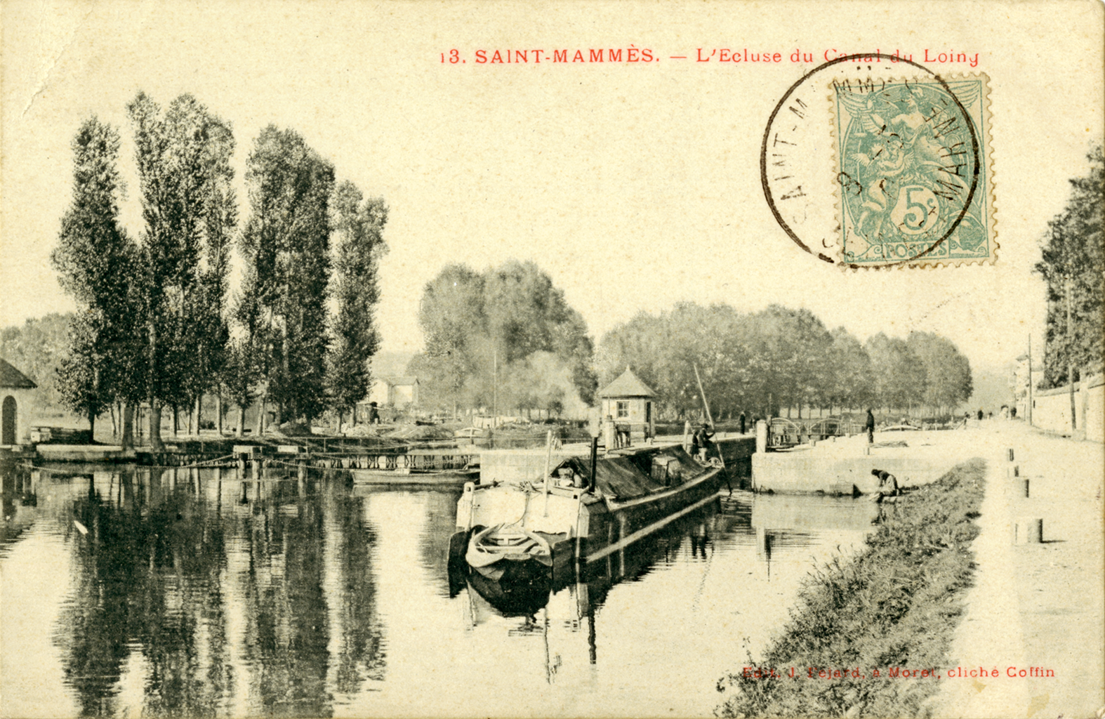 A black and white image of a dock on a river lined with trees and a walking trail. The path is near a small grass shore. A boat with boxes and tarps waits at the dock. On the image is a green stamp, and above it writes &ldquo;3. Saint-Mammès. - L’Ecluse du Canal du Loing&rdquo;