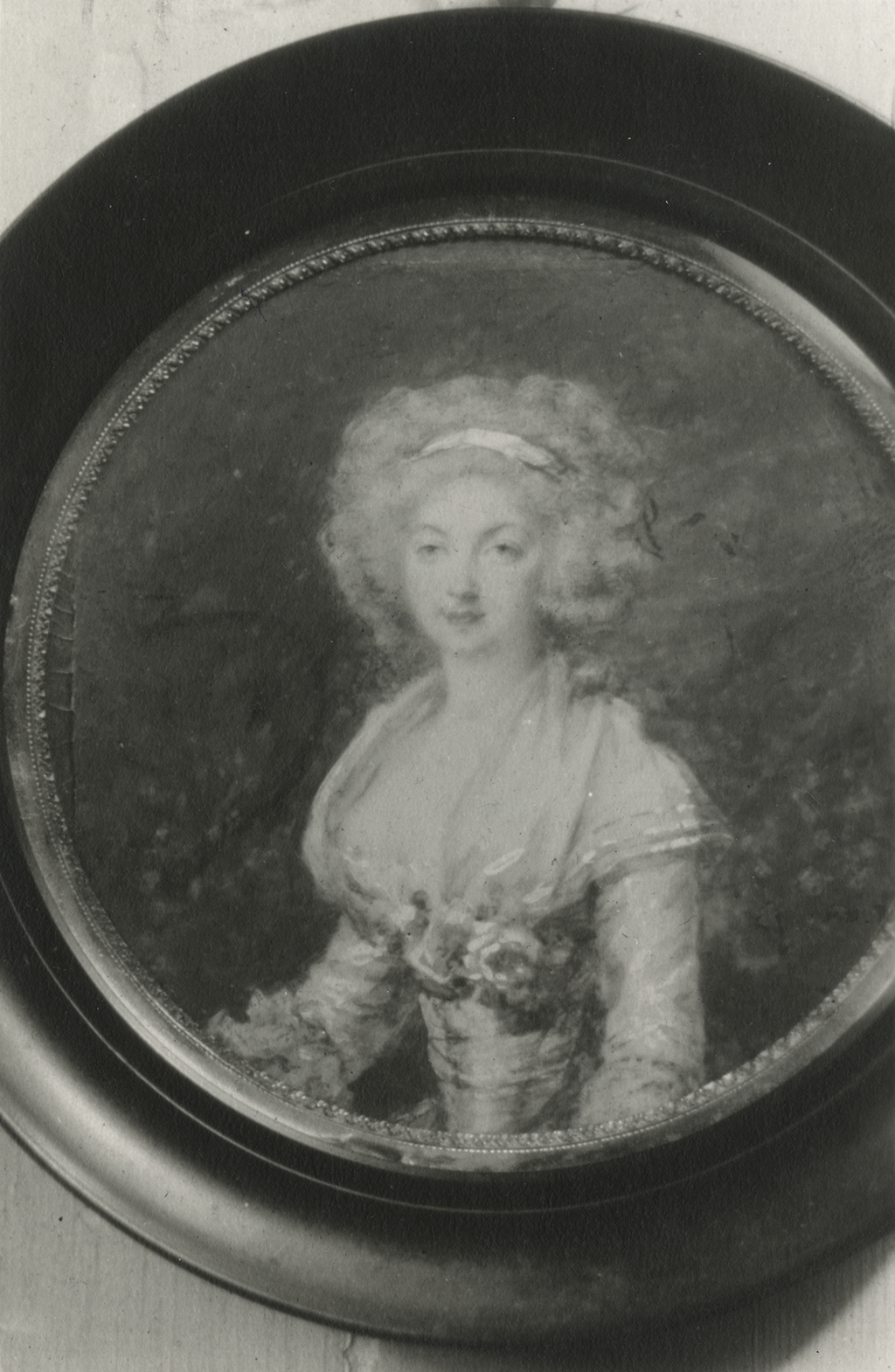 A black and white photograph of a framed miniature painting of a woman. She is shown from the waist up and has large curly hair tied with a headband. She wears a dress with a cloth that covers her shoulder and flowers at her bosom.