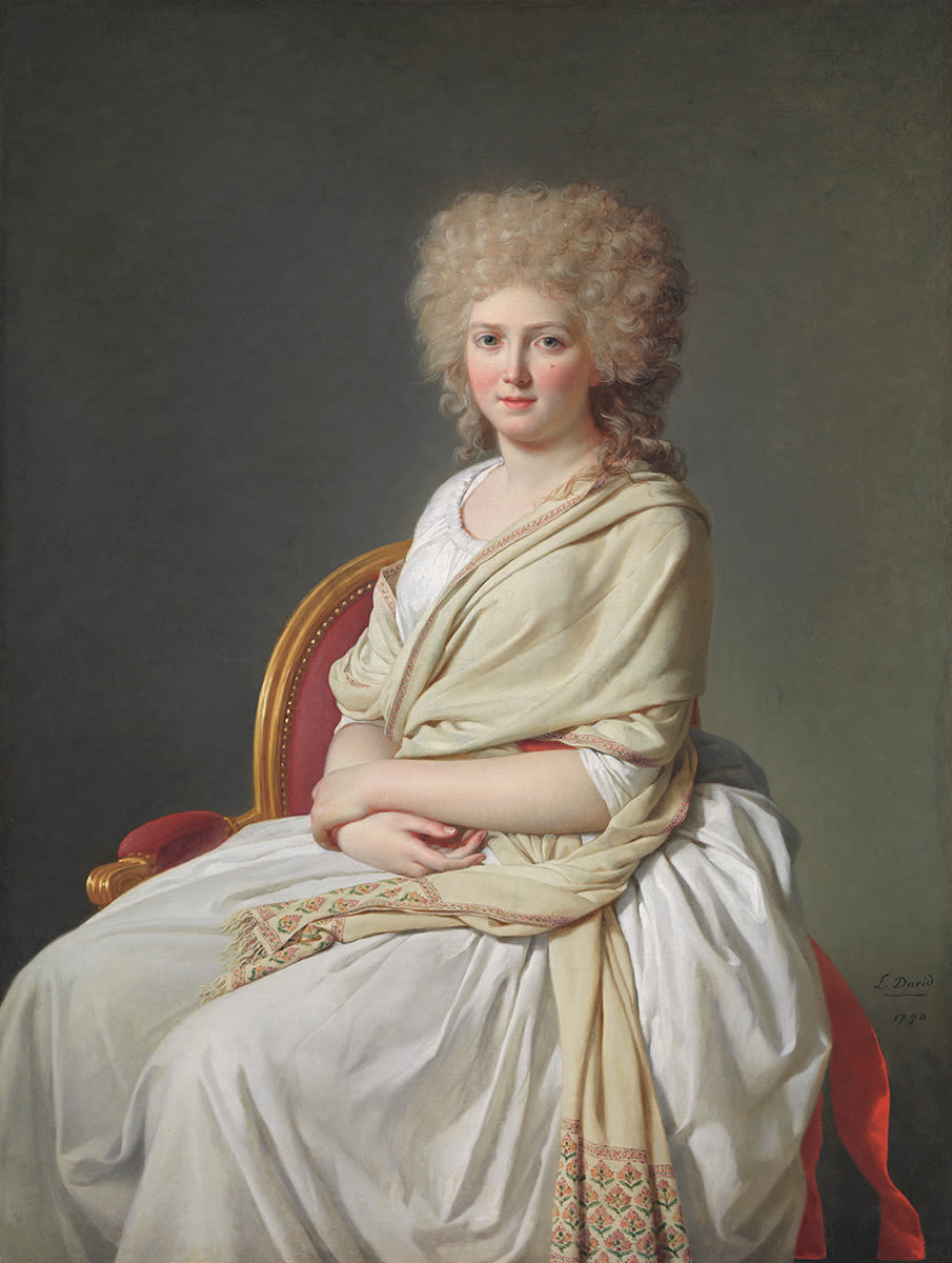 A painting of a light-skinned woman, shown in three-quarter length, wearing a white dress and seated on a golden chair with red upholstery. The woman has green eyes, flushed cheeks, a beauty mark under her left eye, and curly, powdered hair. Over her left shoulder, she wears a long beige shawl that wraps around her chest and has a red and green design embroidered at its ends. She rests both her hands gently in her lap.