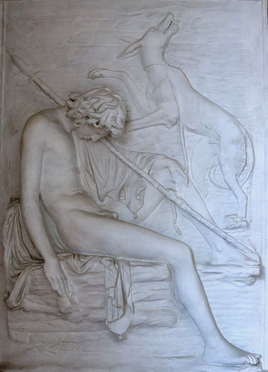 A photograph of a white sculpture depicting a nude man sitting on a rock structure. His head is facing the ground, and he has a spear resting on his shoulder. Behind him a four-legged canine which seems to be climb on top of him.