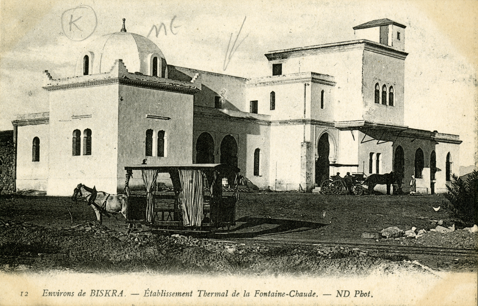 A black and white photograph of a large building with horses and carriages surrounding it. The structure has multiple large with multiple archways floors along with windows on each side.  Under the image it reads &ldquo;Environs de Biskra. – Establishment Thermal de la Fontaine Claude. - ND Phot.&rdquo;
