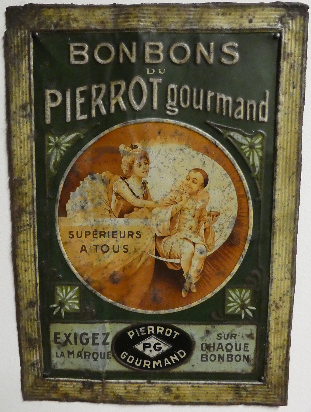 A rectangle cutout of metal with an image that shows a woman holding a child while he sits on a crescent moon with a face. The text at the top states &ldquo;BONBONS DU PIEEROT gourmand&rdquo; The text at the bottom says &ldquo;EXIGEZ LA MARQUE - SUR OHAQUE BONBON&rdquo;