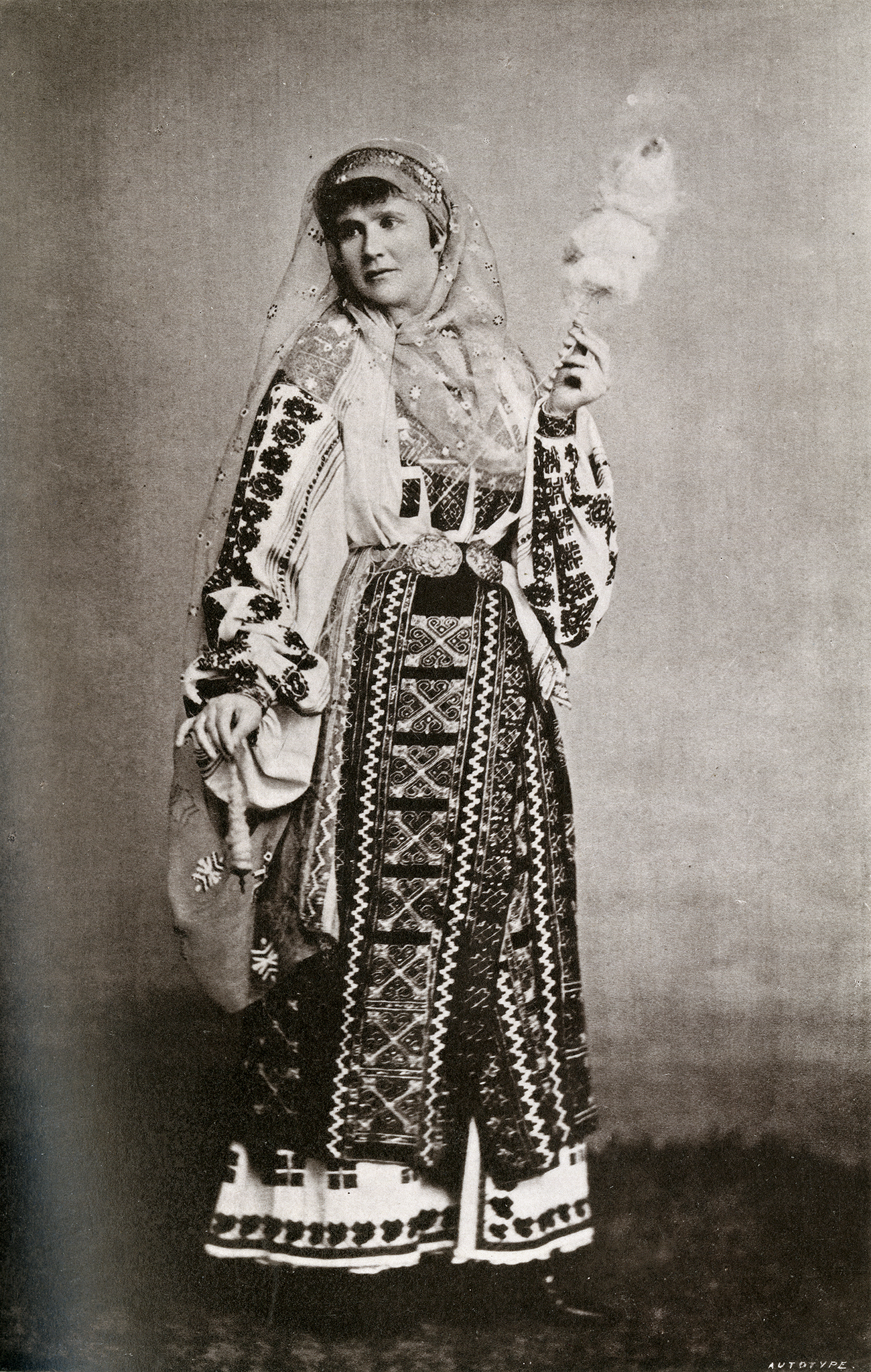 A black and white photograph of a woman wearing a patterned dress along with a hood. She holds a wooden handle that is surrounded by what looks to be cotton.