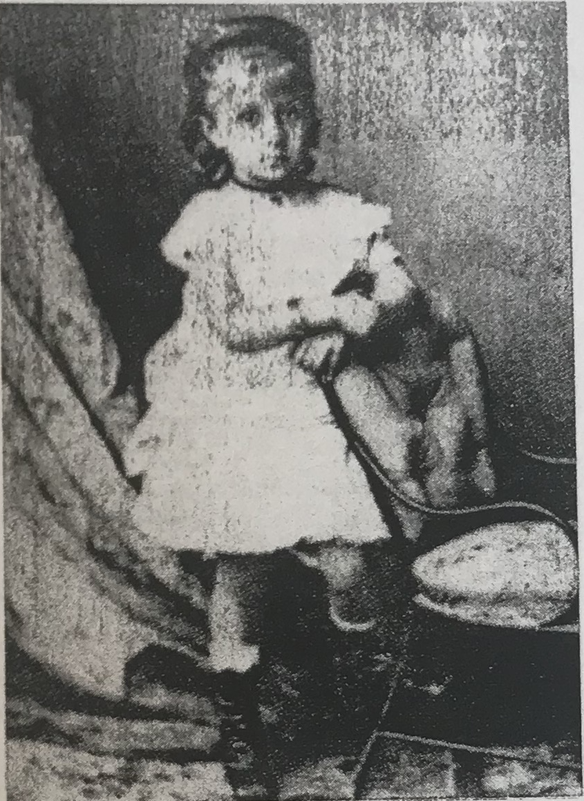 A black and white photograph of a young girl wearing a white dress. She leans against the back of a chair.