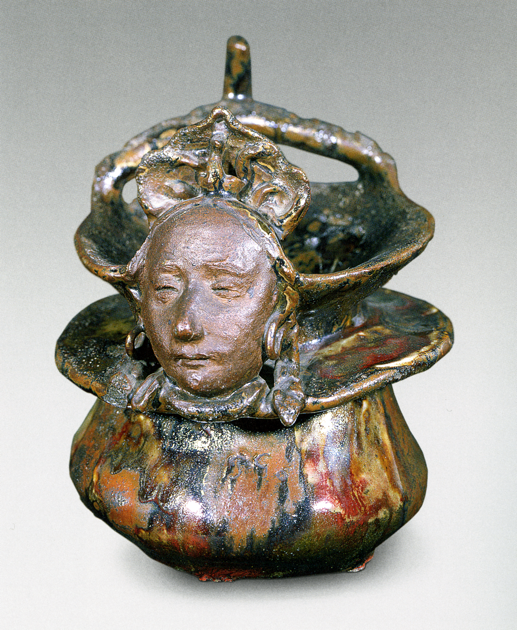 A photograph of a vase with a woman’s head sculpted into it. She appears to be wearing hooped earrings. The vase itself has a mix of various reflective colors such as red, black, and gold or copper.