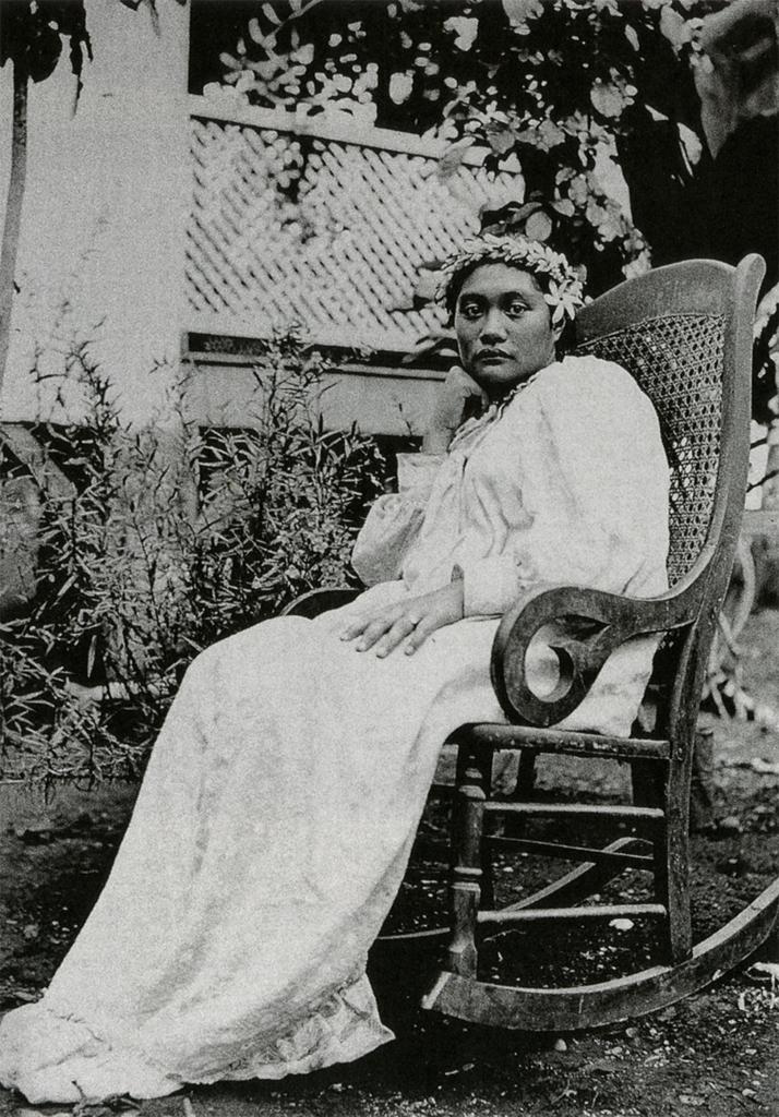 A black and white photograph of a woman white a white dress sitting in a wooden rocking chair. She wears a headband made of flowers.