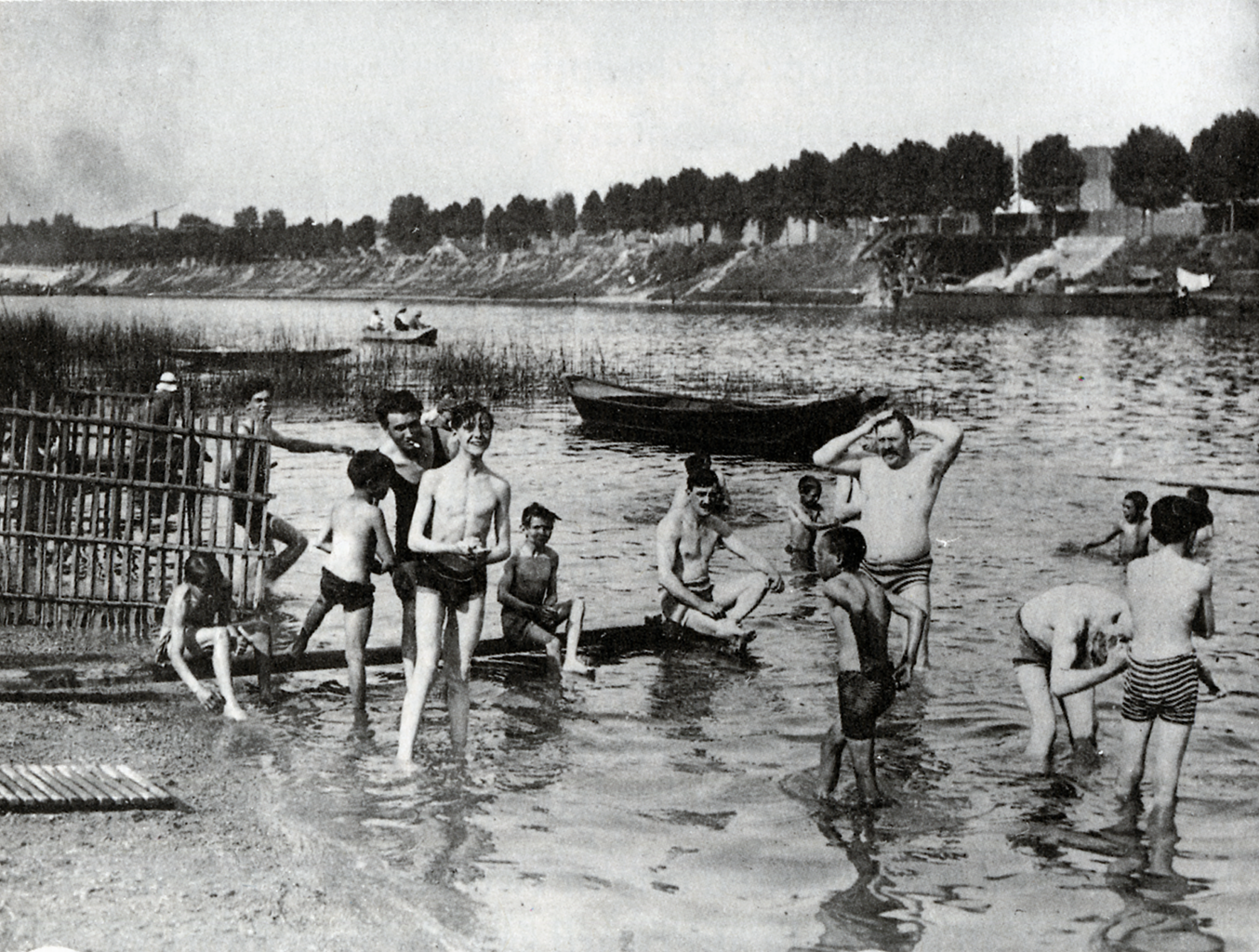 A black and white photograph of a group of men and boys washing themselves in the shallow water near a river. In the background there is a line of trees against a wall.