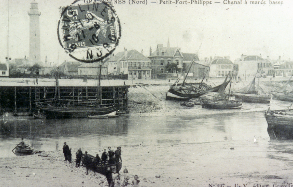 A black and white photograph of a group of people standing on the shore standing by a small river. Wooden boats rest on the banks near the dock. Behind them is a town towered by a lighthouse.  On the top of the photograph is a stamp which has a woman with her arms stretched out on it. At the top of the photograph it says, &ldquo;GRAVELINES (Nord) – Petit-Fort-Philippe – Chenal à màrée basse&rdquo;