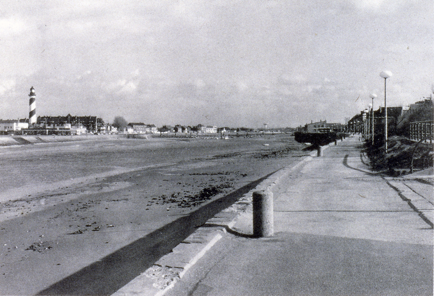 A black and white photograph of a fully evaporated river. To the right, there is a trail with barriers and tall lamps aligned up along it. To the left there is a lighthouse which overlooks a town.