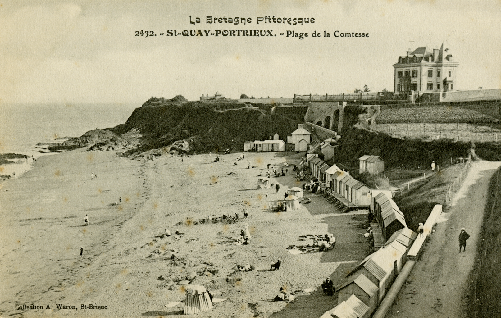 A black and white photograph showing people scattered across the shore of a beach surrounded by small bathing cabins. On the right side of the photograph ther is a road leading to the top of the cliff which rests a white manor. Written at the top of the image is &ldquo;La Bretagne Pittoresque 2432. ~St~QUAY~PORTIEUX. ~ Plage de la Comtesse.&rdquo;
