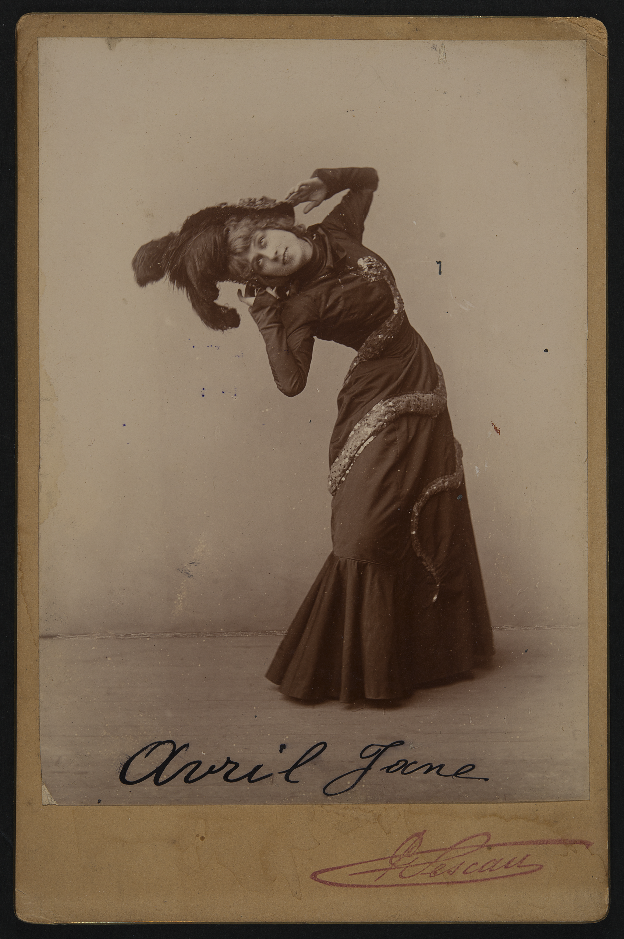 Sepia photograph of a woman bending her back and having her hands near her ears. She wears a dark colored dress and headdress. The bottom of the ground has a signature with &ldquo;Avril Jane.&rdquo;